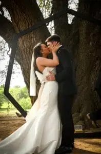 A bride and groom kiss outdoors in front of a large tree and a geometric black frame. The bride wears a white dress and veil; the groom is in a black suit. Sunlight filters through the leaves in the background.