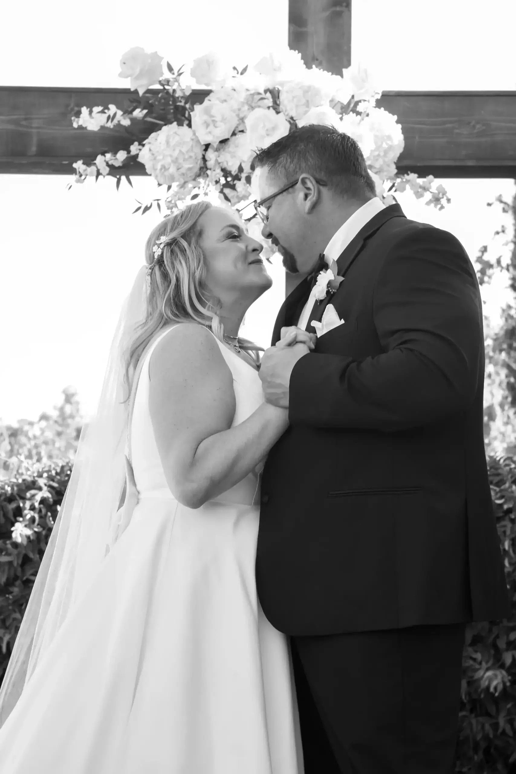 A bride and groom smile and gaze lovingly at each other while holding hands during their wedding, standing outdoors beneath a floral arrangement and wooden cross.