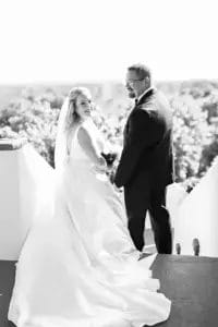 A bride and groom stand outdoors on white steps, holding hands and smiling back at the camera. The bride wears a long white gown and veil; the groom wears a dark suit. Trees are visible in the bright background.