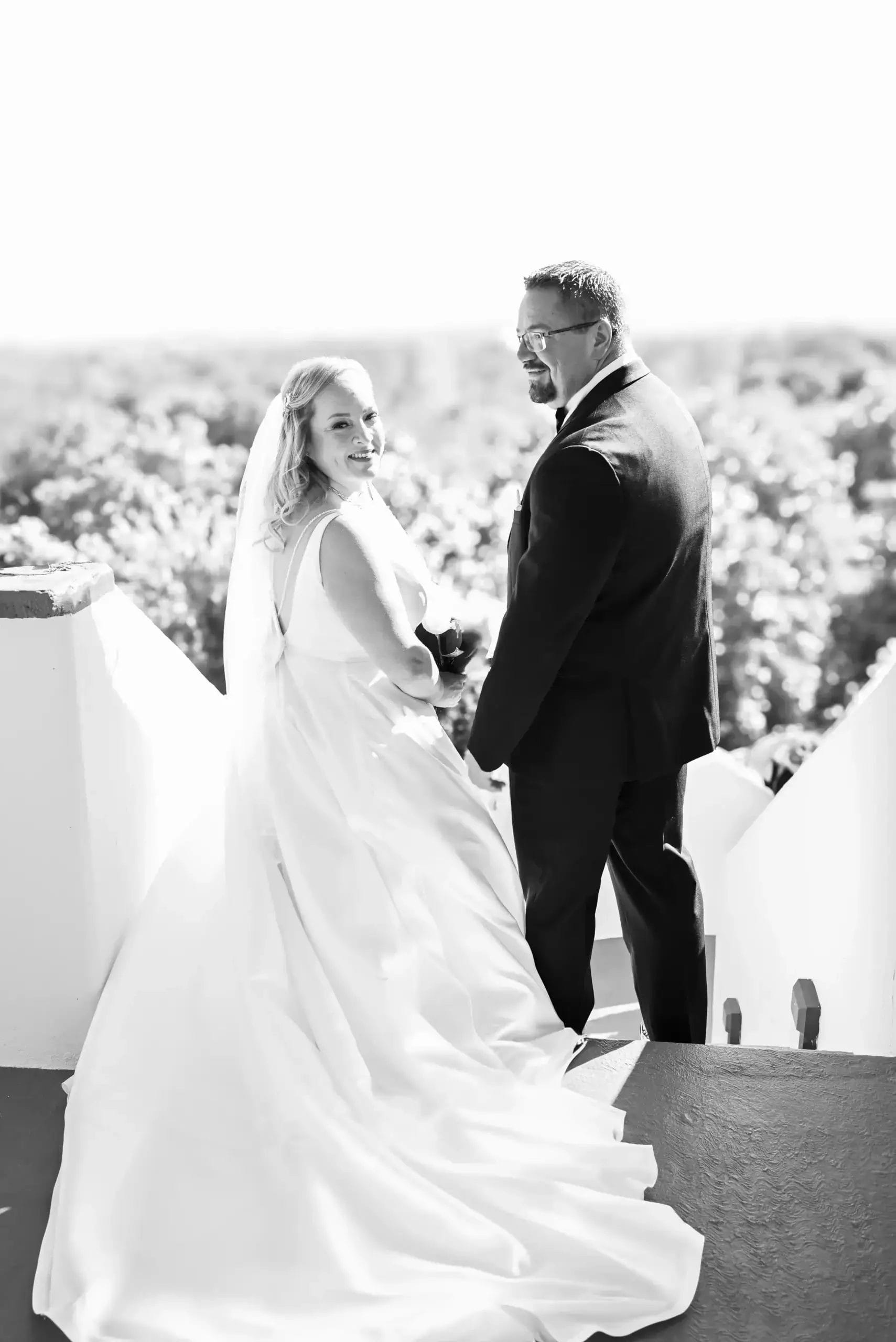 A bride and groom stand outdoors on white steps, holding hands and smiling back at the camera. The bride wears a long white gown and veil; the groom wears a dark suit. Trees are visible in the bright background.