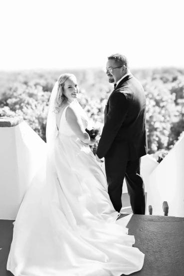 A bride and groom stand outdoors on white steps, holding hands and smiling back at the camera. The bride wears a long white gown and veil; the groom wears a dark suit. Trees are visible in the bright background.