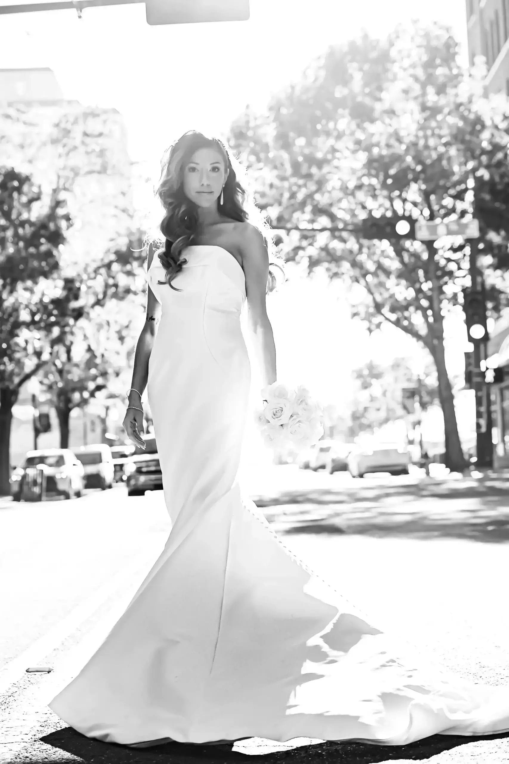 A woman in a strapless, fitted wedding gown stands in the middle of a sunlit city street, holding a bouquet of flowers. Trees, cars, and buildings are visible in the bright background.