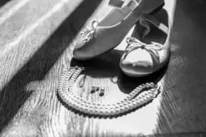A pair of ballet flats with bows, a string of pearls, and a pair of earrings are arranged on a wooden floor, illuminated by sunlight. The image is in black and white.