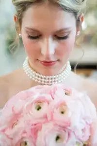 A woman with pearl earrings and a double-stranded pearl necklace looks down at a bouquet of light pink flowers, with soft curls framing her face and natural makeup.