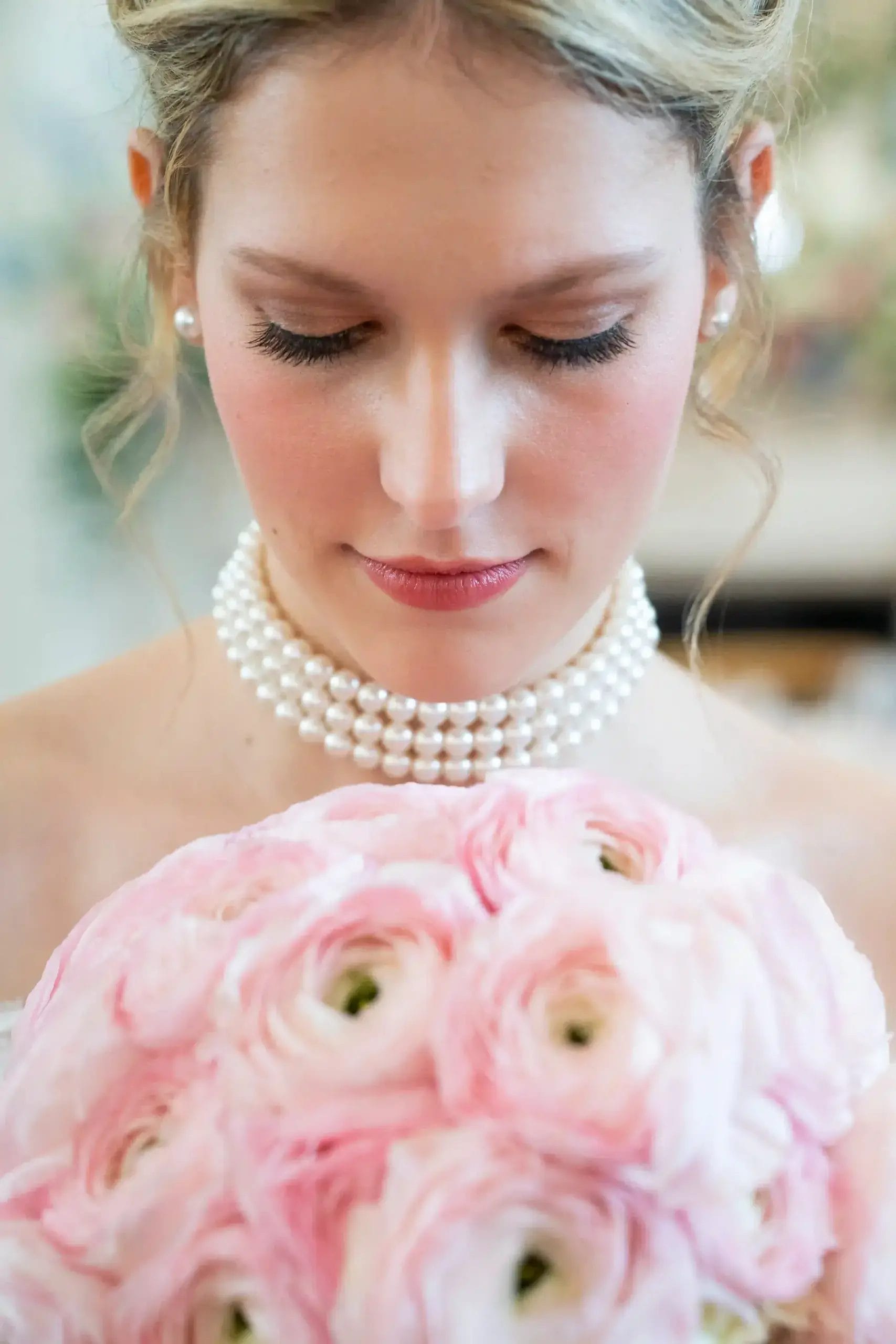 A woman with pearl earrings and a double-stranded pearl necklace looks down at a bouquet of light pink flowers, with soft curls framing her face and natural makeup.