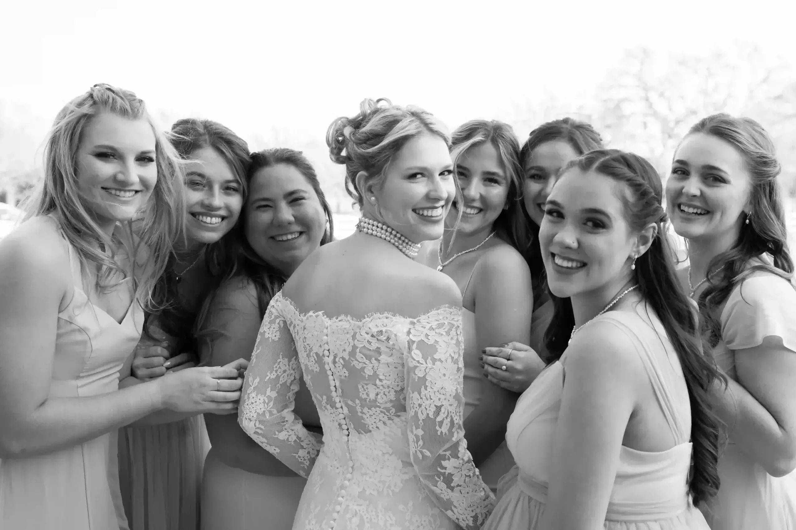 A bride in a lace wedding dress stands smiling and surrounded by six bridesmaids in light dresses, all looking happily at the camera.