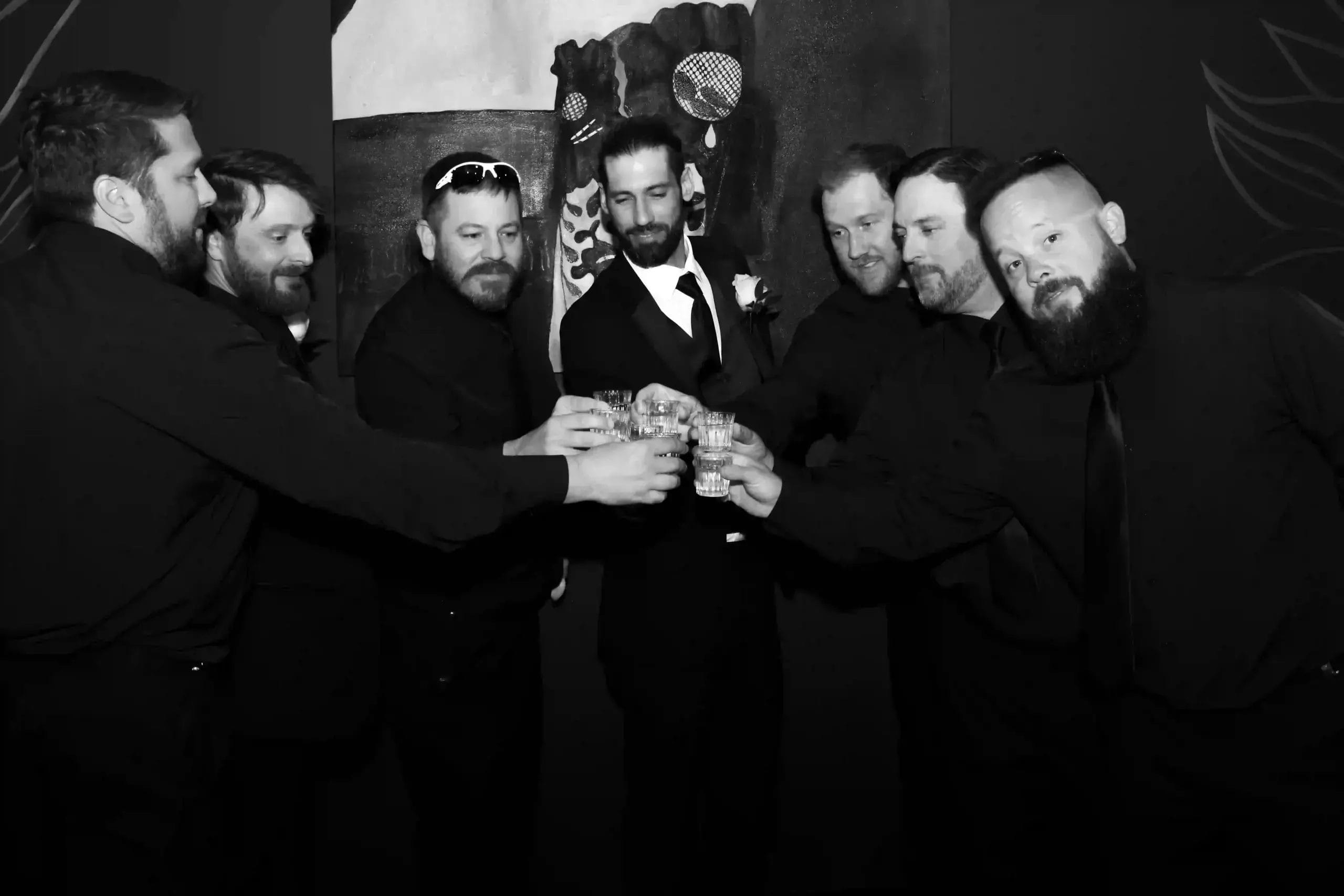 Seven men dressed in dark suits, including one in a lighter suit and tie, stand in a group making a toast with drinks, smiling and facing the camera in a black and white photo.