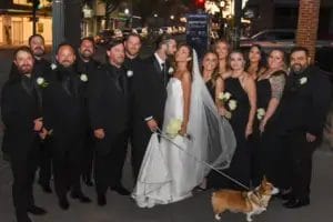 A bride and groom kiss while surrounded by their wedding party dressed in black. The bride holds a bouquet and a leash of a corgi dog. The group smiles on a city sidewalk at night.