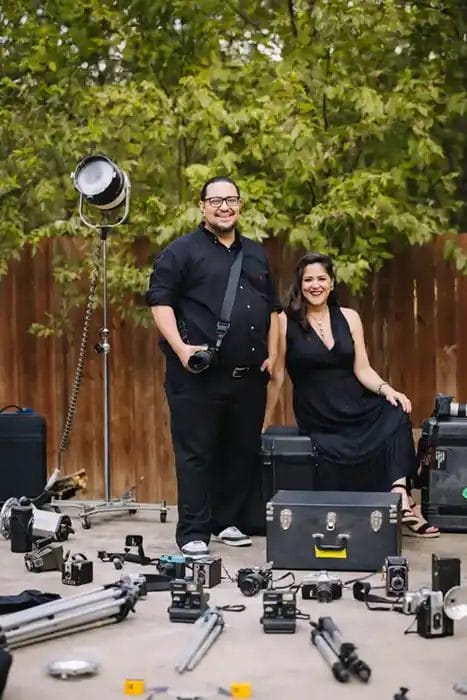 A man and woman dressed in black pose outdoors with vintage cameras, cases, and photography equipment spread out on the ground in front of them. Trees and a wooden fence are in the background.